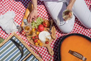Top view of unrecognizable young woman in white pants outside having picnic, eating and playing guitar