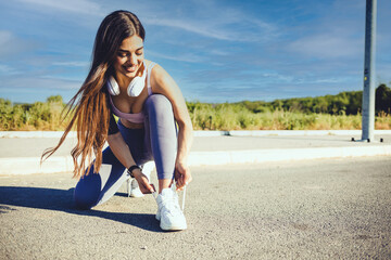 Young fitness woman tying shoelaces on trail. Close up of young woman tying her laces before a run. A fit and active young woman tying her shoes before jogging in a park. - obrazy, fototapety, plakaty