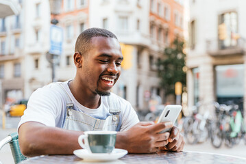 Cheerful young African American male millennial in casual clothes. He is smiling happily while reading good news on smartphone sitting at table with cup of coffee in street cafe