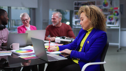 Stylish woman typing on laptop working in creative office with colleagues talking on background