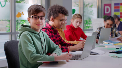 Happy teen boy smile at camera doing homework in classroom using laptop