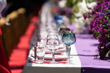 Festive table setting. Crystal and glass goblets of different color on purple white tablecloth. Decor with fresh flowers. Expensive interiors for luxurious wedding or birthday dinner. Selective focus 