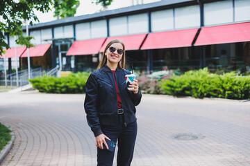 Beautiful and young girl in sunglasses writes a message on a cell phone and drinking coffee outdoors