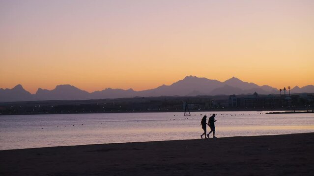 Lovely panoramic landscape natural scene with sea ocean beach a romantic people couple walking around the sand coast with beautiful colorful sunset sky, mountainous on the background. Calm summer day 