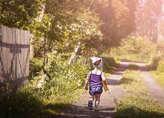 a little girl in a T-shirt, shorts and a white scarf walks with her back to us along a sandy road along a fence, around green trees, grass and flowers