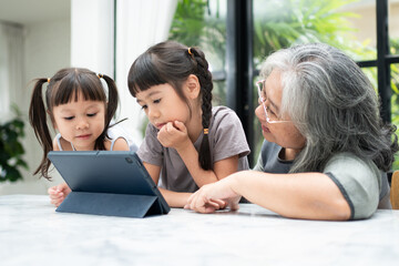Fototapeta na wymiar Asian Grandmother with her two grandchildren having fun and playing education games online with a digital tablet at home in the living room. Concept of online education and caring from parents.
