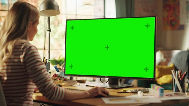 Over the Shoulder: Creative Young Woman Sitting at Her Desk Using Desktop Computer with Mock-up Green Screen. Female Caucasian Specialist Working on Computer with Chroma Key Display at Home