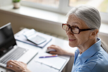 Senior woman using laptop at home. 70 years old lady in eyeglasses smiling at camera, selective focus.