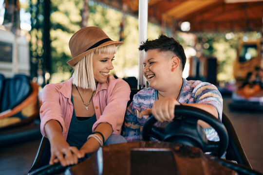 Happy lesbian couple talks while driving bumper car at amusement park.