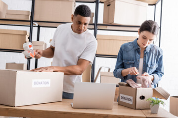 Interracial sellers packaging boxes with donation lettering near laptop in online web store.