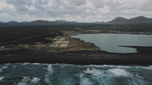 Black Volcanic Sand Beach With Volcanos In The Background Drone Speed Ramp Zoom Out With Ocean Waves At Sunset And Salt Flats Around Ocean Lake Lagoon. Lanzarote Timanfaya National Park.