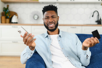 Confused puzzled multiracial man in smart casual shirt holding smartphone and credit card while expressing misunderstanding, spreads his hands in incomprehension - obrazy, fototapety, plakaty