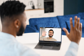 Fototapeta na wymiar Waist up view of the young man in blue shirt waving with hand and saying hello while chatting via laptop with male colleague from home