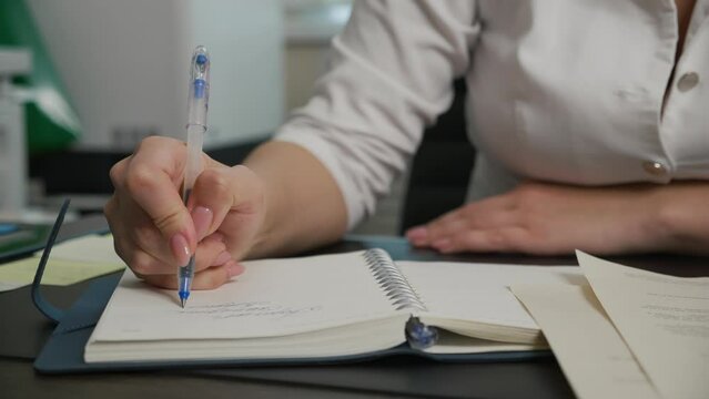 In medical office cabinet young female doctor in open white lab coat with rolled up sleeves sits at table with documents on desk writing cursive words in notebook with blue ink pen, slow motion pan.