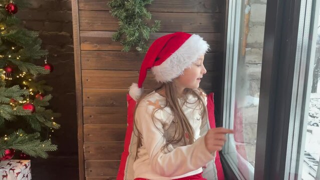 a little Christmas girl at the window, a happy girl in a Santa hat and New Year's pajamas breathes on the window and draws a picture on it with her finger