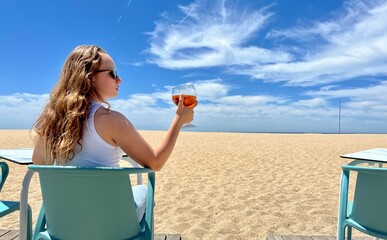 a teenage girl is sitting on the background of the ocean or the sea in her hands she has a glass of iced tea she is in a white T-shirt her hair is loose with her back to the camera . High quality