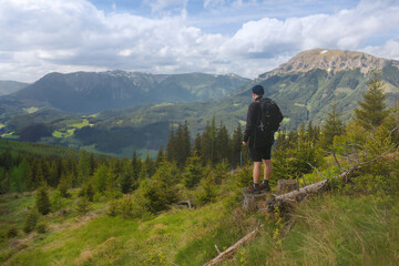 A lone man with a backpack on his back stands on a nature trail in the mountain landscape of Austria, with the Schneealpe and Raxalpe mountain ranges visible in the background.