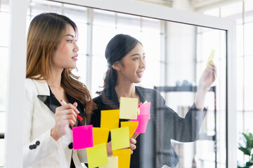 Group of successful Asian businesswomen teamwork. Brainstorm meeting with colorful sticky paper notes on the glass wall for new ideas. Using agile methodology for business in a tech start-up office.