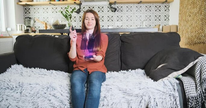 Woman sitting on a sofa in the living room chooses shoes through a hologram on a smartphone screen. 3d shoes are spinning over the phone screen, the hand changes shoe models with a wave. Metaverse