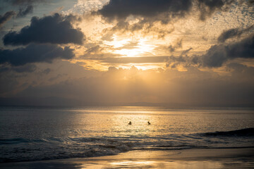 Fototapeta premium silhouette shot of people swimming under the golden sunlight god rays with clouds surrounding the sun and the blue gold water with waves lapping the shore showing peaceful life at havelock andaman isl