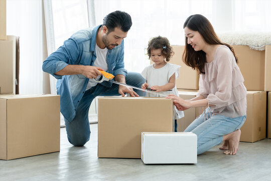 Mixed race family, Latino father and Asian mother, with little cute daughter using scotch tape for packing cardboard boxes on the floor. Young family preparing stuff for move to new house together