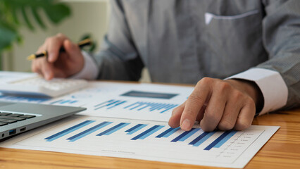 Businessman hand holding pen pointing to marketing graph and working on laptop computer and financial documents Concept of online accounting work in the office