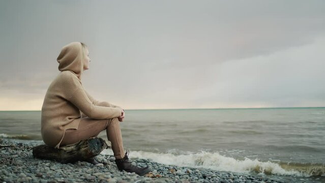 Side view: A woman in a warm sweater with a hood on her head sits on the ocean shore