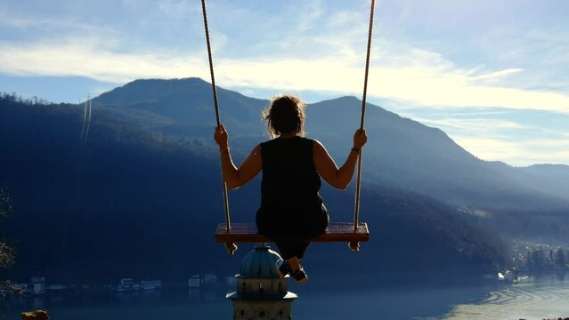Woman Sitting on a Swing with Sunlight and Mountain View with a Church Tower on Lake Lugano in Morcote, Ticino, Switzerland.