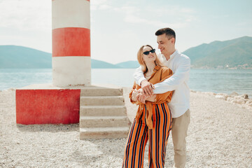 A couple stands near a lighthouse on the beach against the backdrop of the sea and mountains....