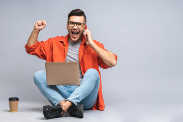 Successful winner! Portrait of a happy young man sitting on the floor, using laptop and celebrating success or victory isolated over white grey background. Using mobile phone.