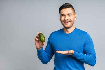 Smiling ukrainian young man holding fresh avocado fruit showing isolated over grey white background...
