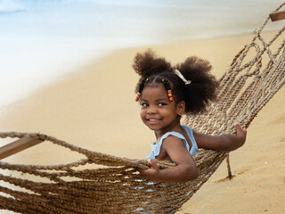 Pretty little African girl giving a big smile while sitting in the rope hammock on the beach. Summer vacation holiday concept.