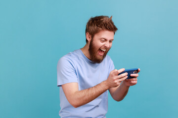 Portrait of handsome young adult bearded man standing, using smartphone and playing mobile game with excited positive face, happy to win. Indoor studio shot isolated on blue background. © khosrork