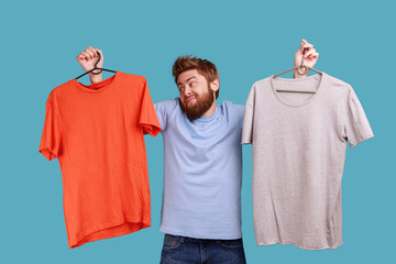 Portrait of puzzled doubtful handsome bearded man holding two hangers with gray and orange T-shirts, choosing outfit for going for a walk. Indoor studio shot isolated on blue background.