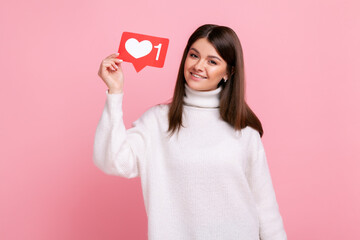 Charming young girl holding heart like icon of social media and looking at camera with toothy smile, wearing white casual style sweater. Indoor studio shot isolated on pink background.