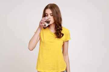 Portrait of attractive thirsty woman of young age with brown wavy hair in yellow casual T-shirt drinking water from glass, water balance. Indoor studio shot isolated on gray background.