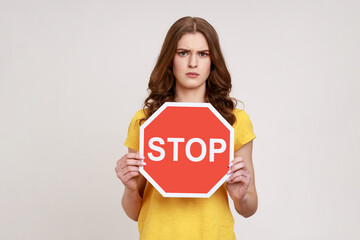 Fototapeta na wymiar Ban, forbidden access! Young serious woman in yellow T-shirt holding red stop sign, warning and looking angrily and strict at camera. Indoor studio shot isolated on gray background.
