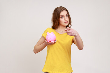 Beautiful woman of young age in yellow T-shirt investing in bitcoins, holding piggy bank and pointing on you with finger, looking at camera with smile. Indoor studio shot isolated on gray background.