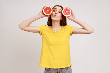 Portrait of positive teenager girl with dark hair holding two half slices of grapefruit with funny expression, making kissing gesture, looking at camera. Indoor studio shot isolated on gray background