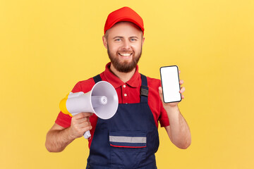 Happy smiling worker man in blue overalls and red cap holding megaphone for making announcement and...