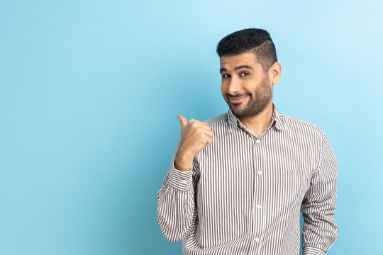 Portrait of pleasant looking bearded man pointing thumb finger aside at copy space, showing area for advertisement, wearing striped shirt. Indoor studio shot isolated on blue background.