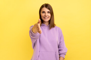 Fototapeta premium Nice to meet you. Portrait of pleased friendly smiling woman stretching her hand welcoming guests, hospitality, wearing purple hoodie. Indoor studio shot isolated on yellow background.