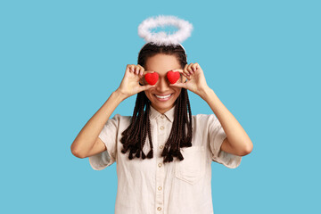 Portrait of woman with black dreadlocks covering her eyes with red hears, having angelic nimb over head, smiling happily, wearing white shirt. Indoor studio shot isolated on blue background.