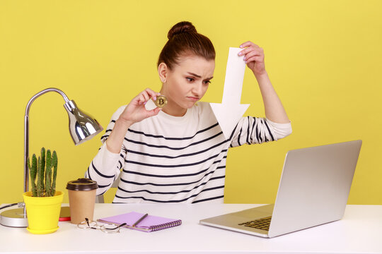 Dissatisfied woman sitting on workplace showing gold bitcoin and arrow pointing down to laptop display, downgrade of electronic currency. Indoor studio studio shot isolated on yellow background. - Powered by Adobe