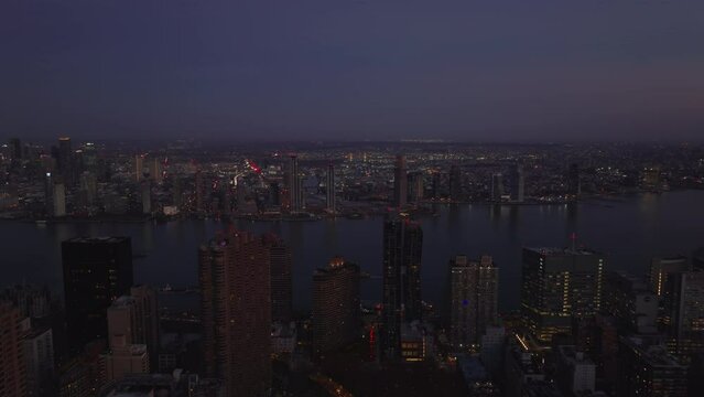 Forwards fly above city after sunset. Aerial view of wide East River surrounded by high rise buildings on waterfronts. Manhattan, New York City, USA