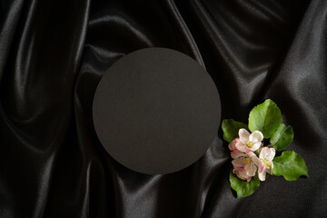 Round geometric platform podium stand for cosmetics product presentation and spring blooming tree branches with white flowers on elegant black silk satin fabric background. Top view