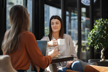 Two young business women in a cafe having one on one meeting. Friends after work talking gossiping and having coffee at a window table on a sunny day..