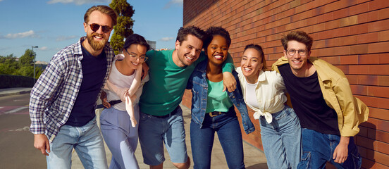 Happy diverse friends enjoying time together. Six different young Caucasian, Afro American and Asian people in fashion clothes standing on street, hugging each other, looking at camera and smiling