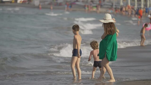 Mother holding baby and two children walk on soft sea sand meeting with waves, family summer vacation