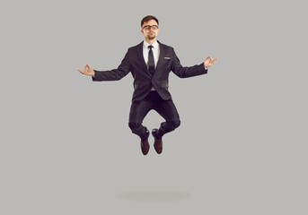 Tranquil male entrepreneur levitating while meditating. Studio portrait of handsome young business man in suit and glasses practising meditation exercise, reaching zen and floating in air in yoga pose - obrazy, fototapety, plakaty